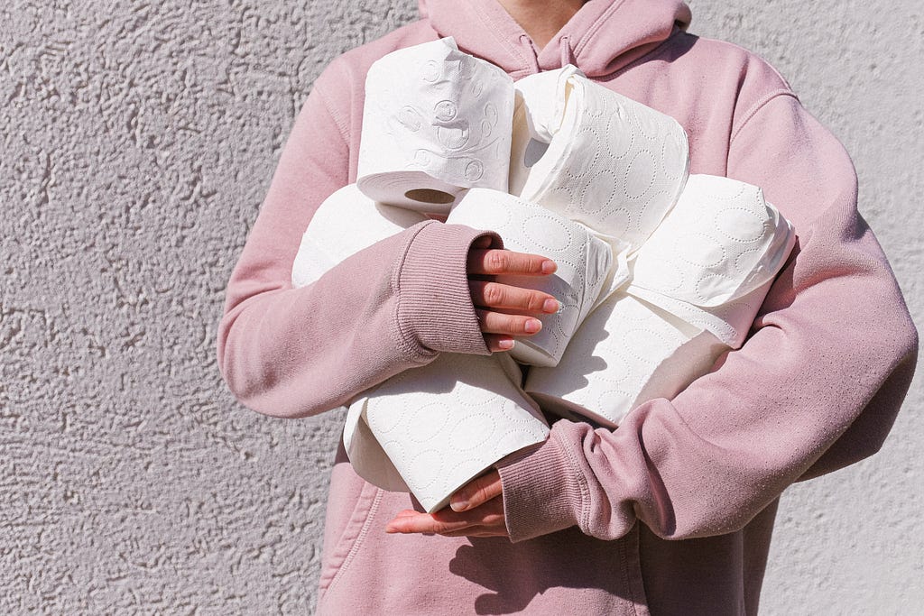 A person in a light pink hoodie holds 7 rolls of toilet paper in their arms.