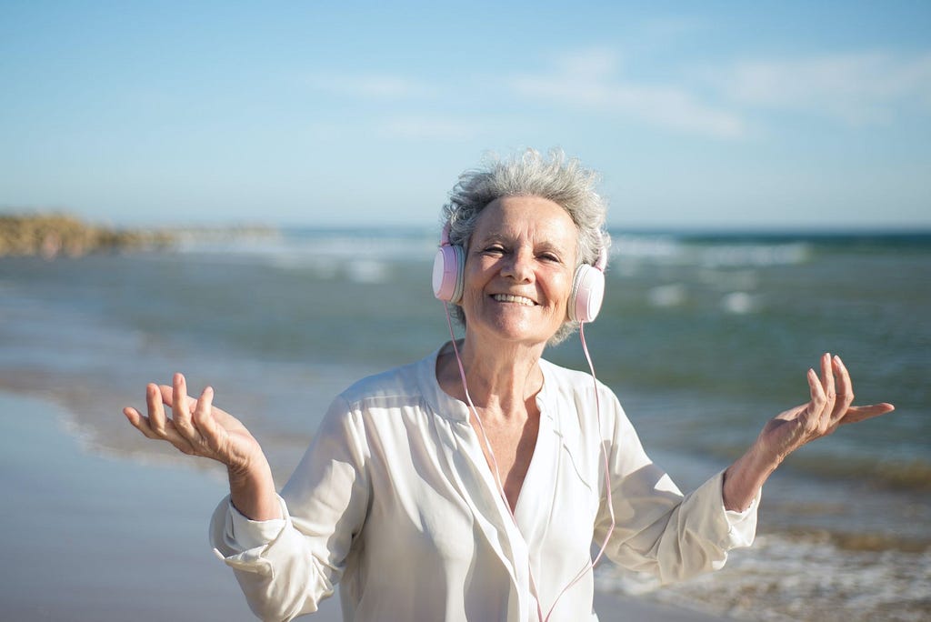 Picture of an older lady standing on a beach in a flowy white shirt with headphones on