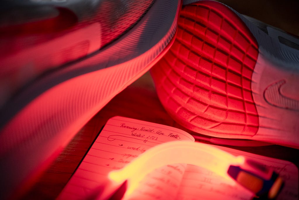 An armband light illuminates a pair of running shoes and the first spread of my fall/winter 2023 training journal, which holds the slow and careful build of mileage I have planned, in red light. Oberhausen, Germany, September 17, 2023.