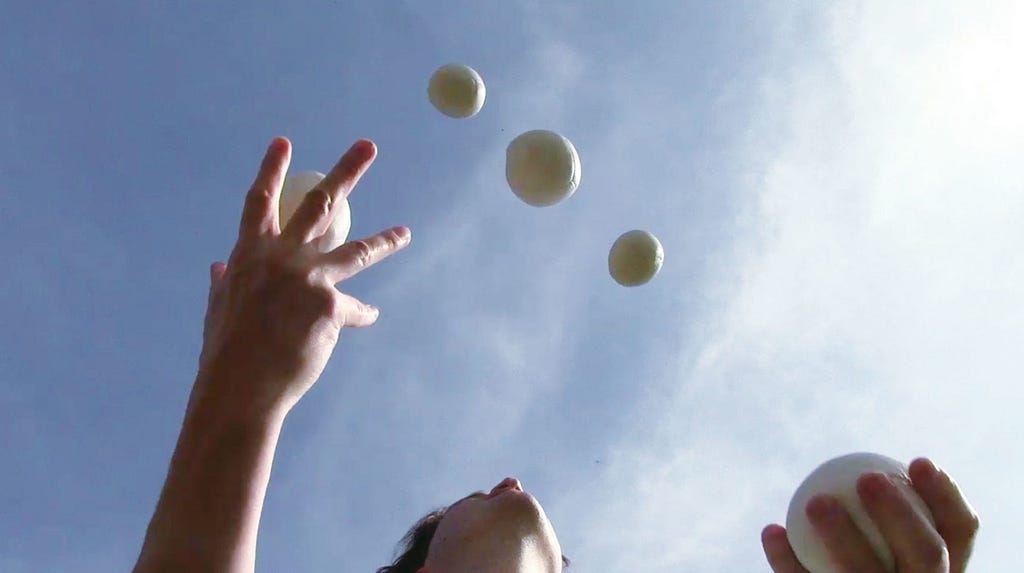 From below, we see a person juggling five white balls against a blue sky