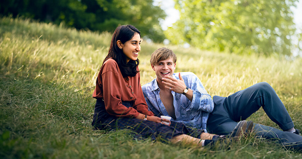 In a field full of grass with blurry trees in the background, two people are relaxing and laughing. The girl has long hair and a red long-sleeved shirt and pants while the guy has blonde hair and wearing an opened blue flannel and jeans.