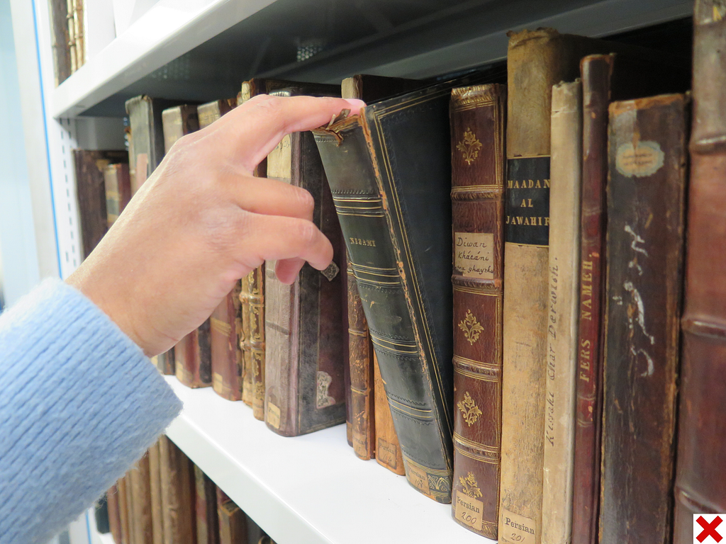 Demonstration of unsafe book removal. A hand grabs the top of the spine of a book in order to pull it out from the shelf, causing visible damage. There is a red cross in the bottom right-hand corner to signal improper practice.