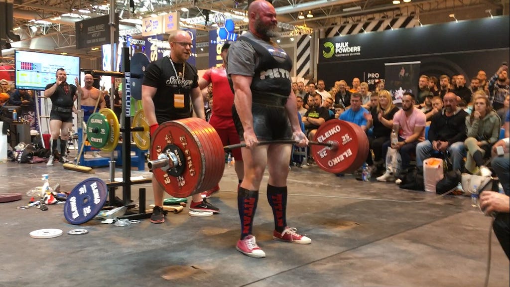 Man holding a heavy barbell at a competition, surrounded by other people cheering