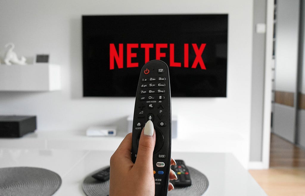 Uma mão segura um controle remoto em frente a uma televisão com a logo do serviço de streaming Netflix.