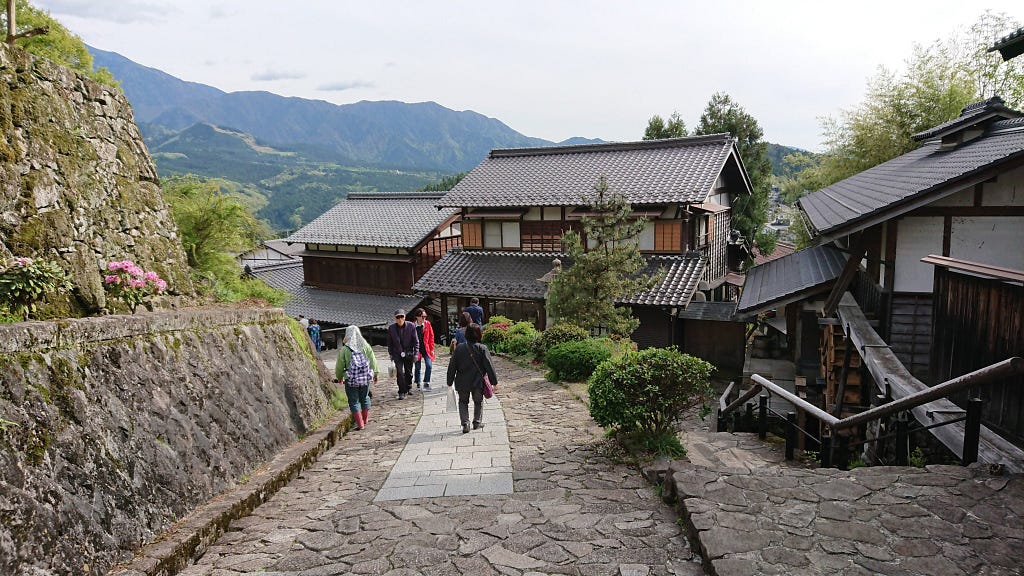 A paved mountain road leading down to the traditional town, with blue mountains in the distance.