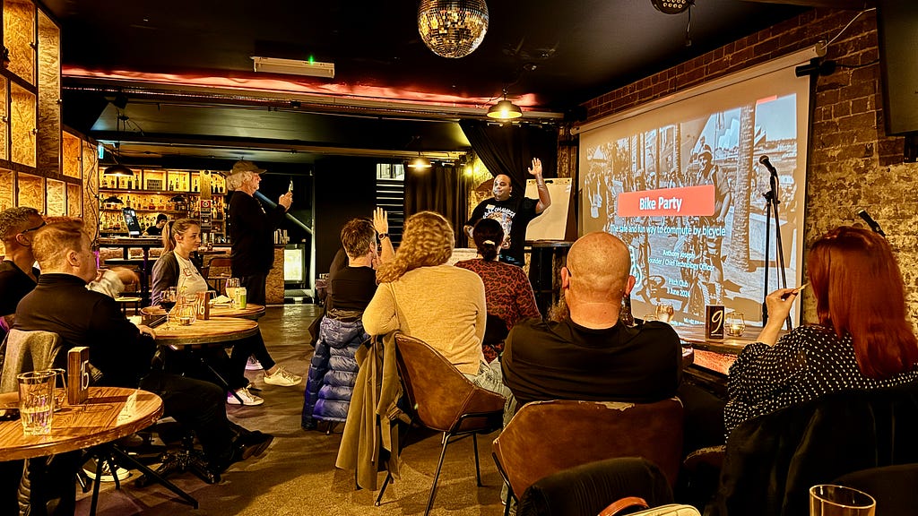 Middle-aged man of south asian appearance in a black t-shirt raising one arm and holding a microphone in the other. Standing in front of a projection screen showing a pitch deck titled “Bike Party”. Several startup founders seated in the audience at low bar tables in small groups. They are in an underground bar where the dim lighting has an orange tint.