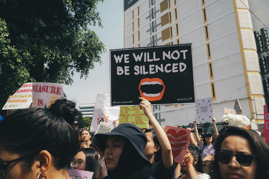 A group of women are protesting in public. There are many billboards and one of them says “we will not be silenced” and shows a drawing of an open mouth.
