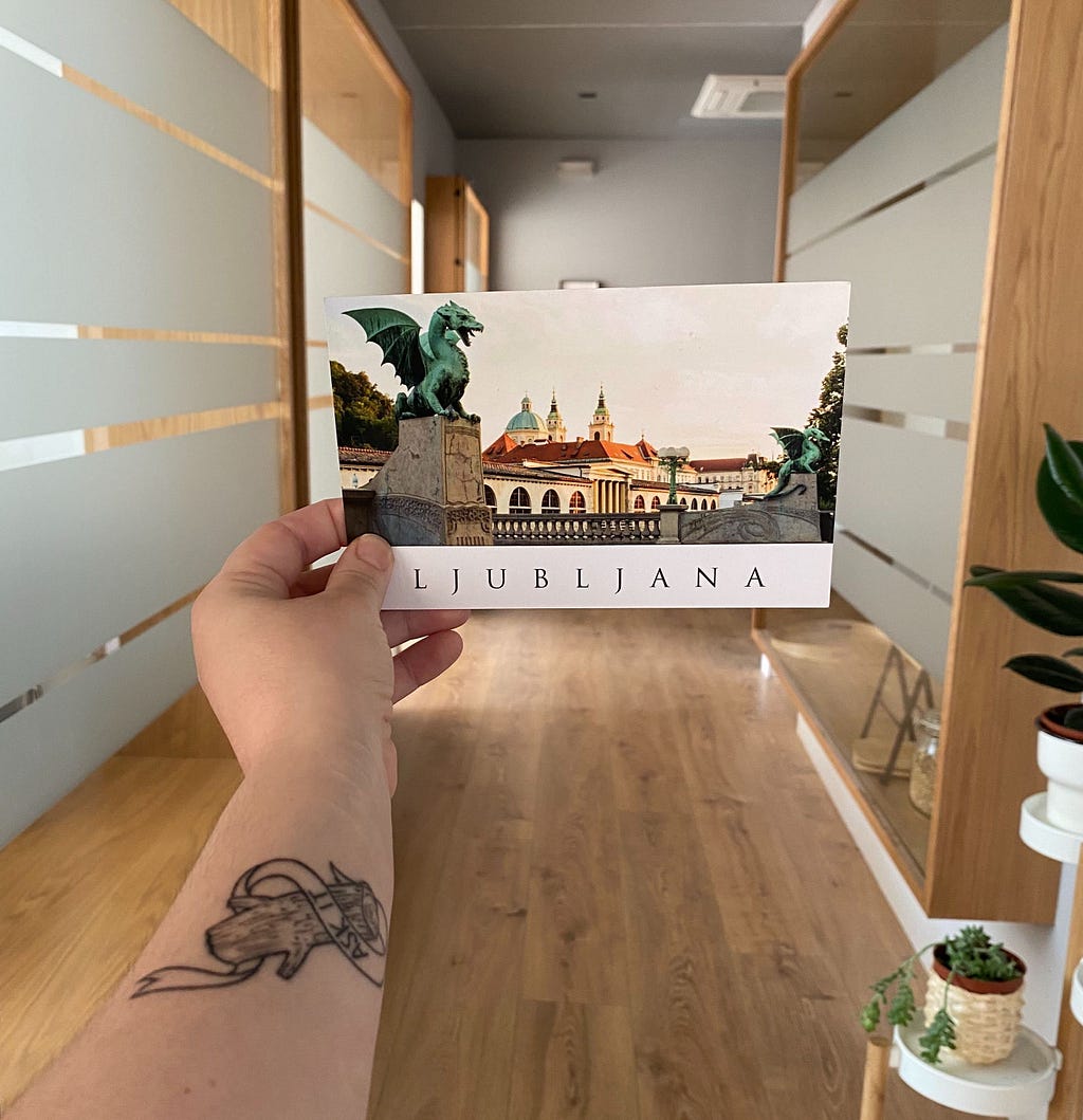 An arm holding a postcard of a dragon statue and building, the postcard reads, “Ljubljana.”