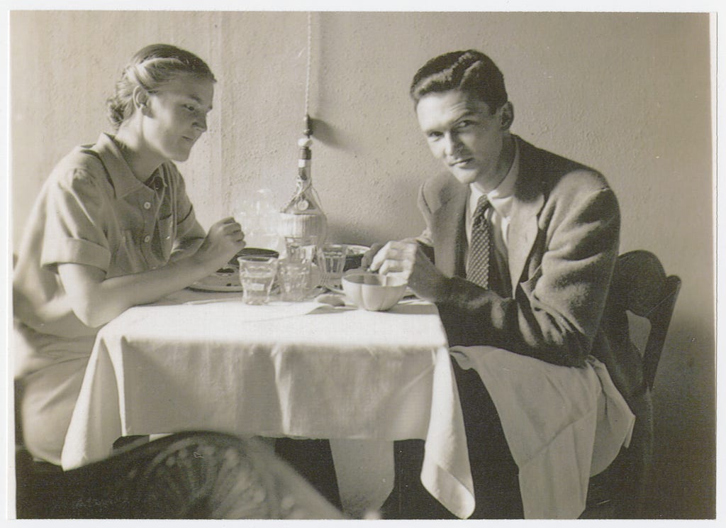 A woman with her hair pulled back in a low bun sits at a restaurant table with a man wearing a suit and tie. There is a white tablecloth on the table. The woman and man lean toward their food.