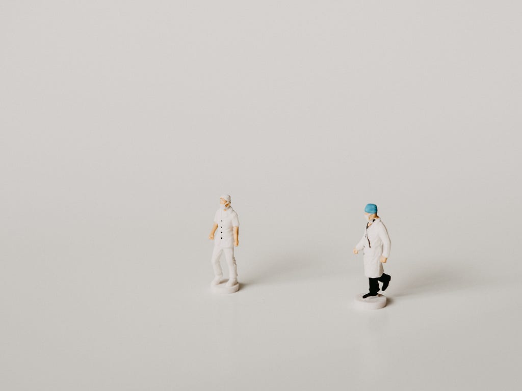 Two miniatures on a white background. One miniature is a nurse in all white scrubs and shoes. The other miniature is a doctor with a blue cap, stethoscope, white lab coat, and black pants and shoes.