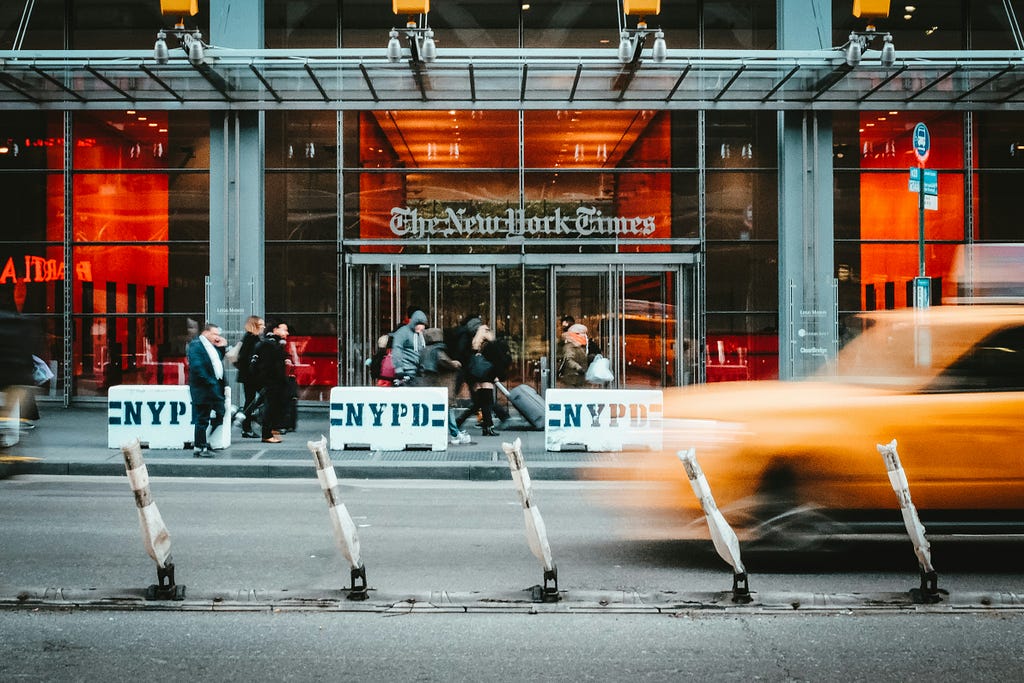 Photo of the entrance to The New York Times building with a blurred yellow cab passign in front