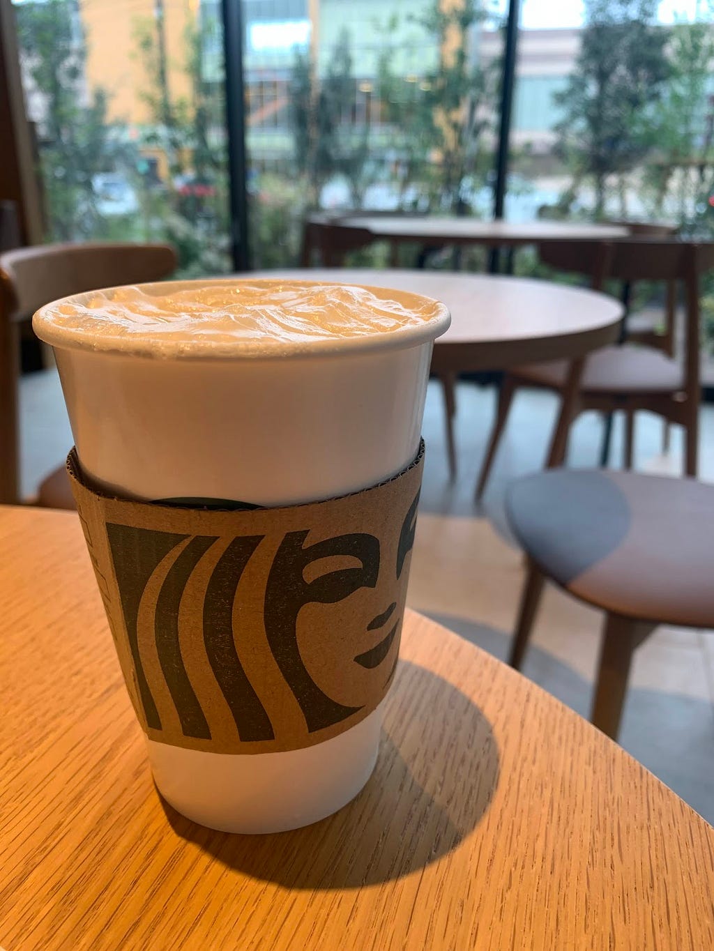 A cup of Starbucks latte on a wooden table inside a cozy café, with a view of the greenery and seating area outside the window.