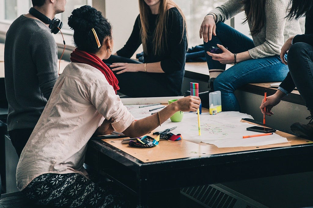 An image of 5 people sat around a desk having a discussion. There’s a big sheet of paper on the desk and lots of pens and drawing equipment.