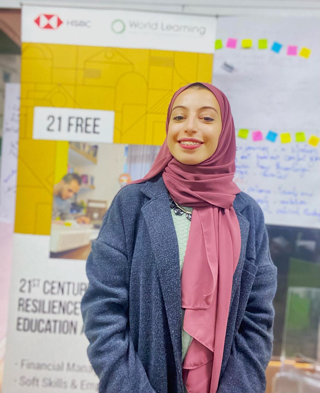 A woman in her 20s wearing a pink hijab is smiling at the camera. Behind her is a tall banner that has World Learning’s logo on it and the words ‘21 FREE’ on one side and a whiteboard with post-it notes.