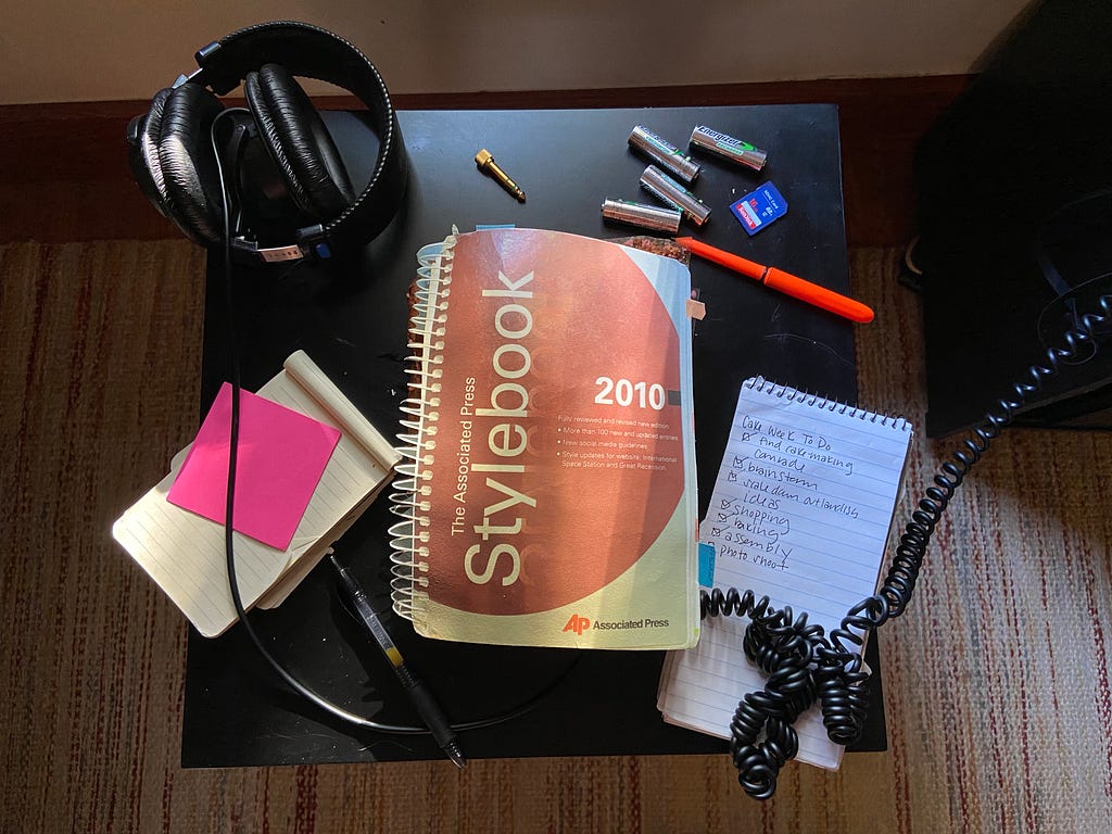 A photo of a cake that looks like a spiral-bound Associated Press Stylebook from 2010. It is on a black table alongside pieces of paper, sticky notes, headphones, and pens.