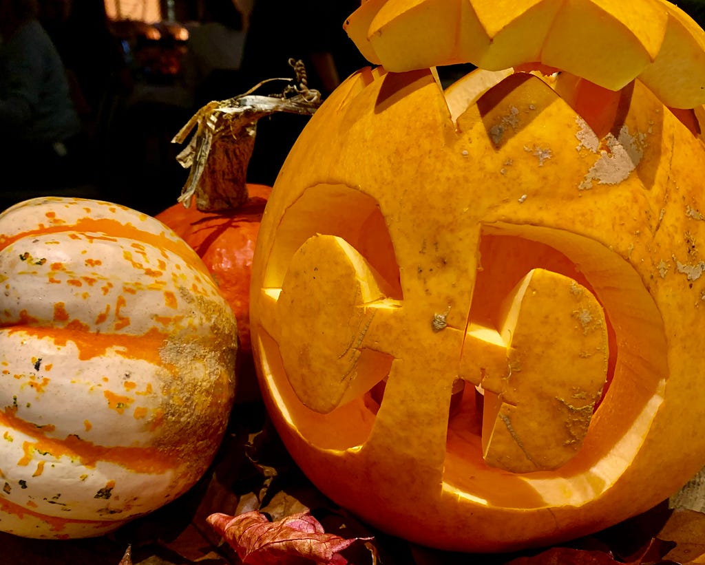 Three pumpkins from a table display. The largest pumpkin has been carved as a jack-o’-lantern with the letters ‘OD’ carved out instead of a typical jack-o’-lantern face.