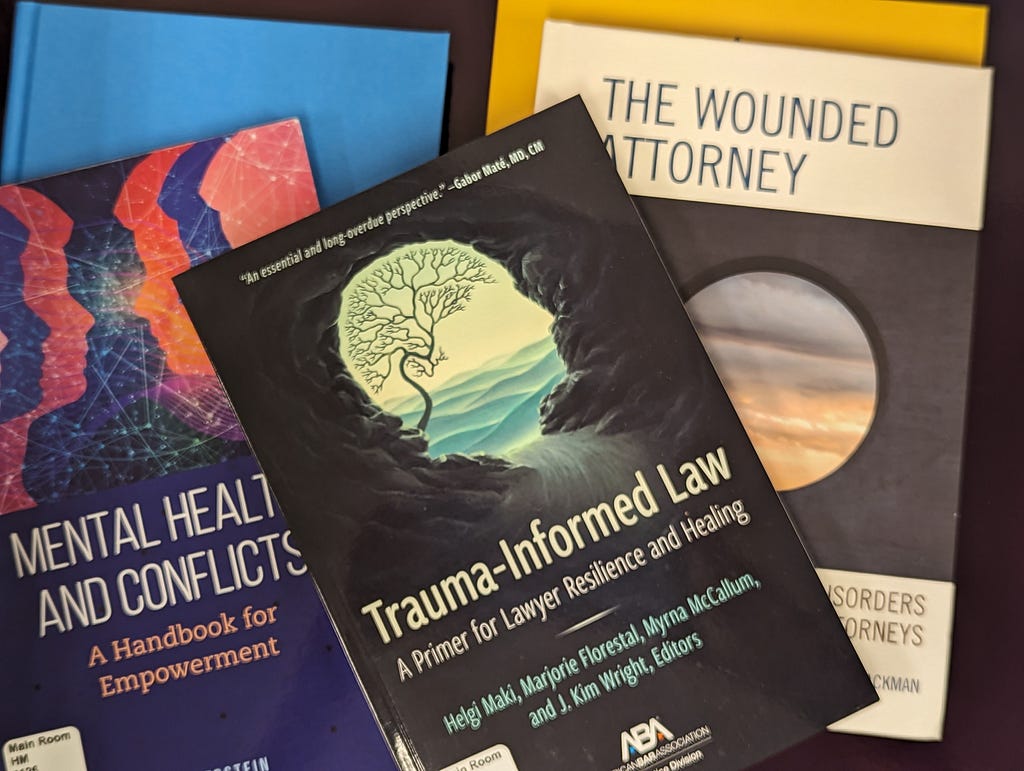 Five books are scattered on a surface. The title of one book is mostly visible and reads, “Mental Health and Conflicts” in white text. An image above the title displays three human faces in profile, and of different colors, facing three other faces, also in profile. Another book has the title “Trauma-Informed Law” printed in cream colored text. The image above the title is of another human face in profile with illustrations of a tree and water superimposed. One additional book title is visible.