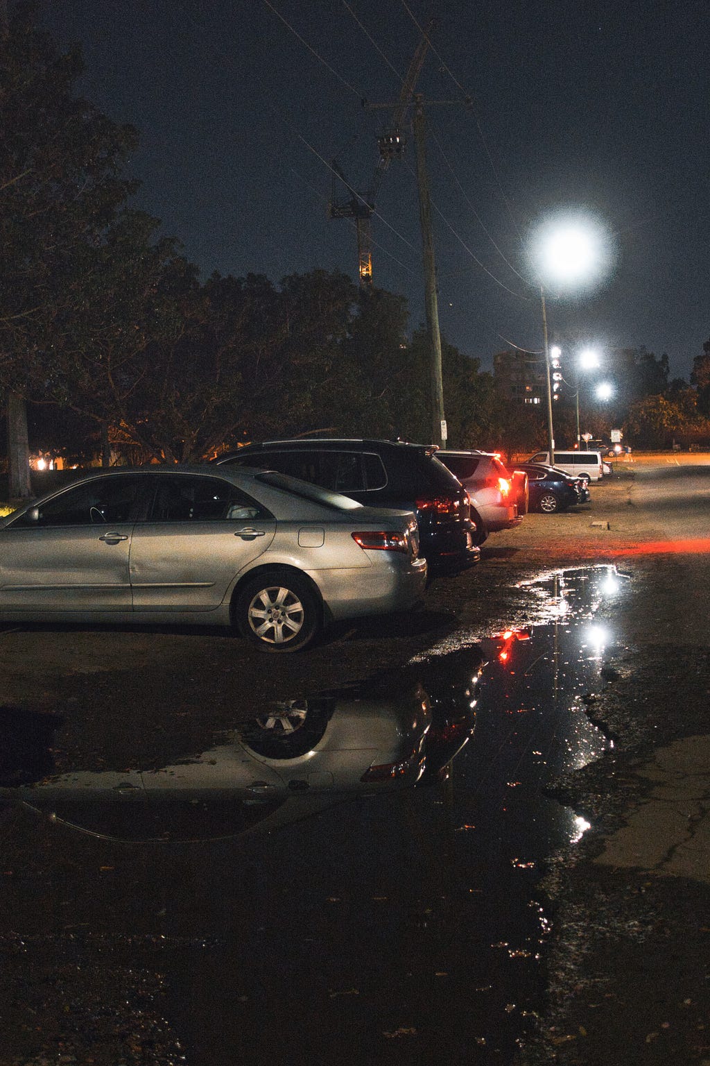 An outdoor dirt parking lot with cars pointing to the left. The puddle in front of the camera reflects the light above and along the street.