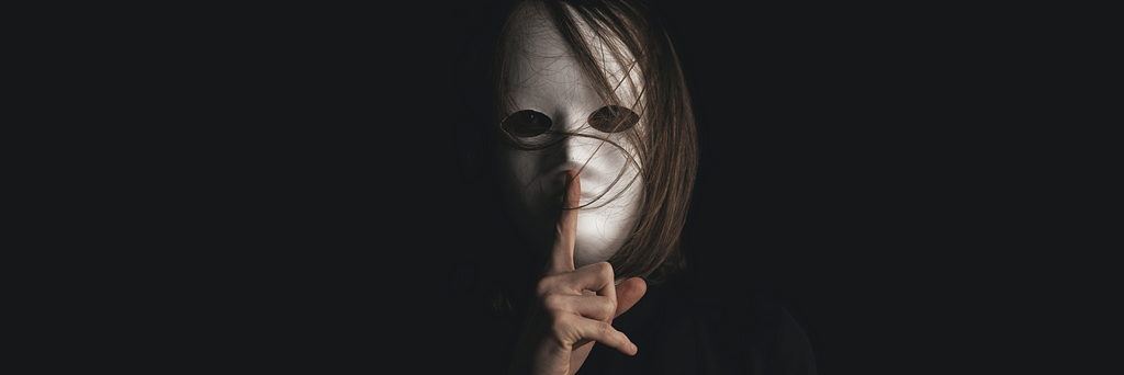Pessoa com cabelos longos, usando uma máscara branca cobrindo seu rosto, fazendo sinal de silêncio com a mão em frente a sua boca
