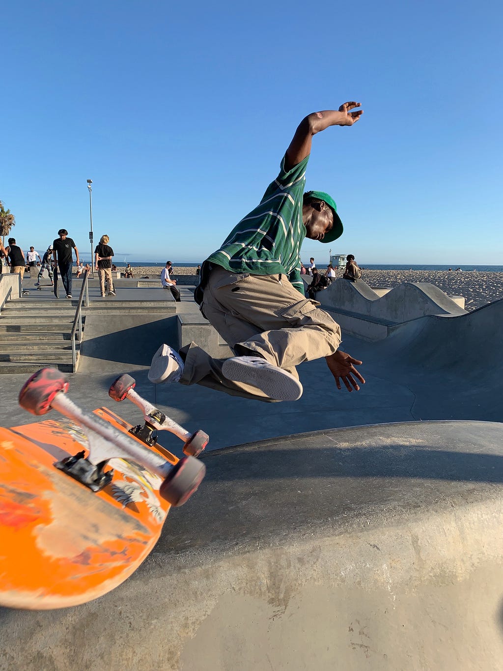 A Black man in a green shirt and khaki pants is pictured mid air as he does a flip on his skateboard at a Venice Beach skate park.