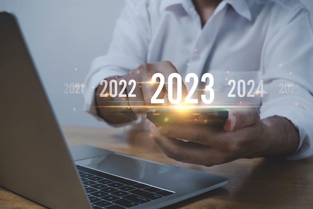 Person on their cell phone and laptop with different years popping out digitally over the screen, and the year “2023” highlighted.