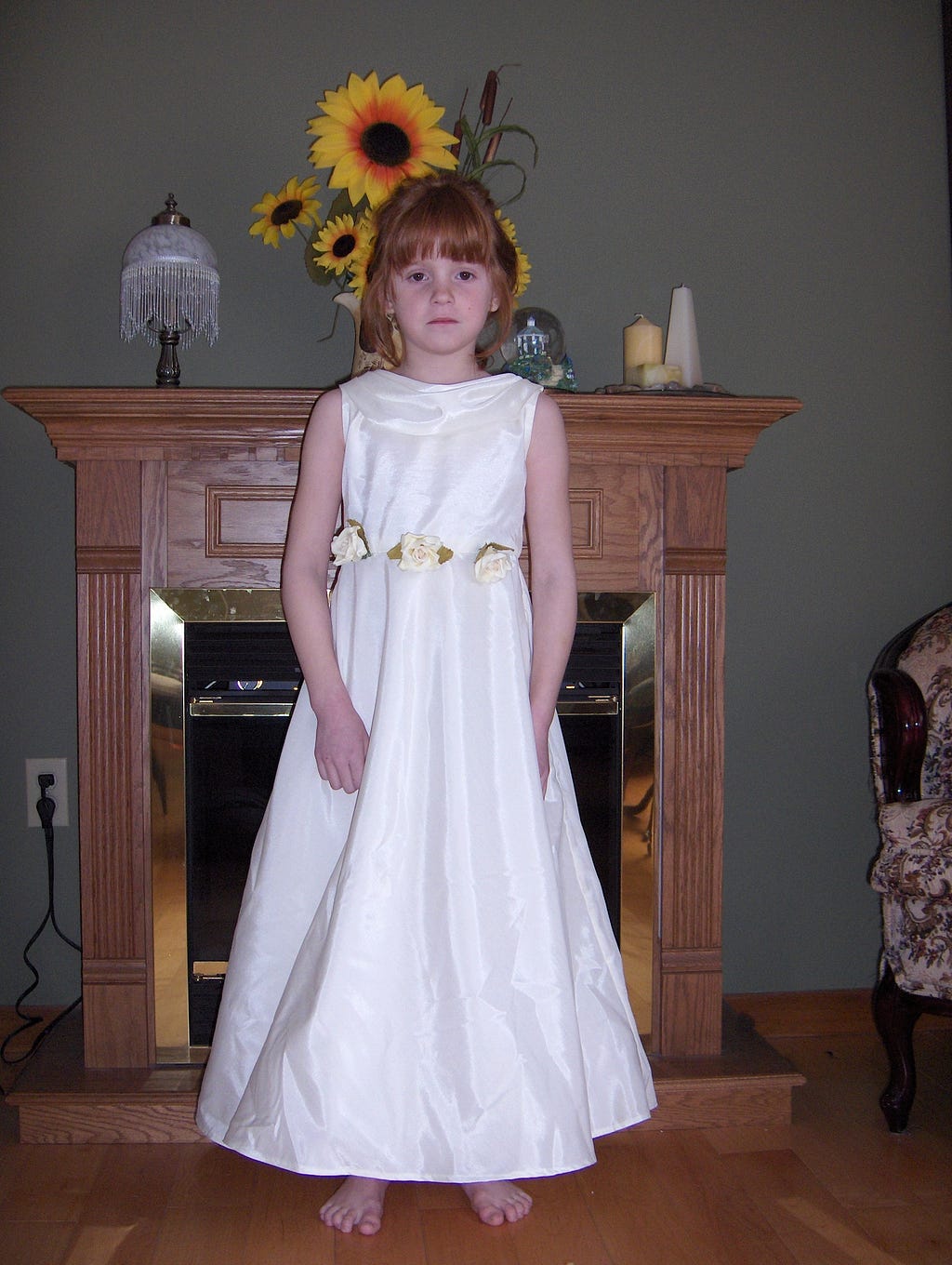 Young Fiona stands in front of a fireplace mantle in a white sleeveless dress. She is not smiling.