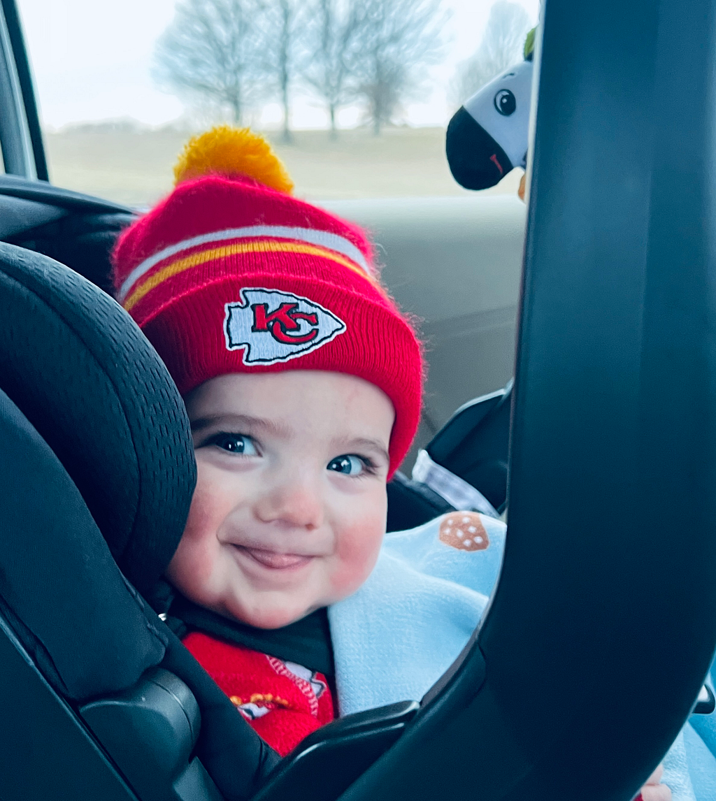 An adorable baby wearing a KC Chiefs hat smiling at the camera from a carseat.