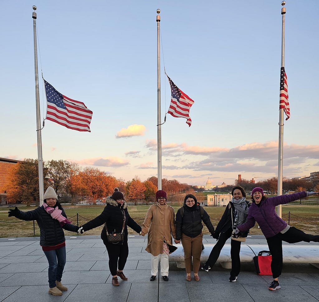 Six people hold hands in front of three American flags waving during a strong wind.