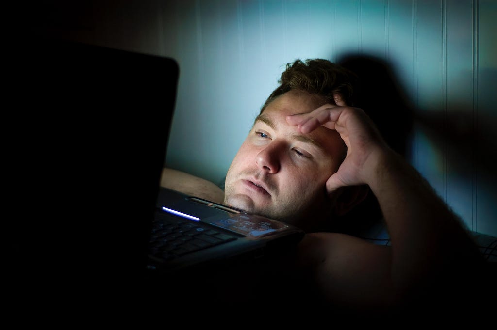 A man lying in bed, staring at a laptop screen