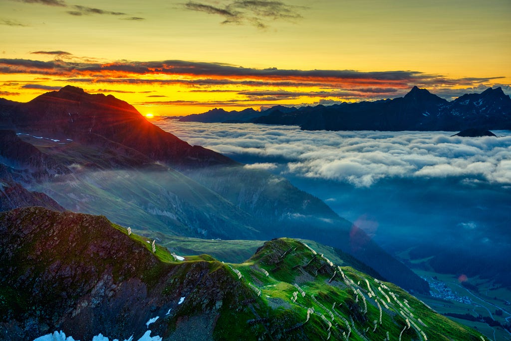 Brightly colored picture of the sun rising above mountain peaks and clouds. The sky is golden and there is a green field on top of the mountain in the foreground.