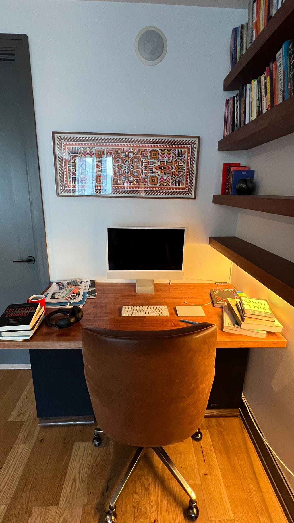 Photo of Austin’s desk. There is a small wooden desktop with an Apple monitor and keyboard, with short stacks of books on both sides of the table and a pair of over ear headphones. On the wall above the desk there is an intricate geometric illustration in various colors.
