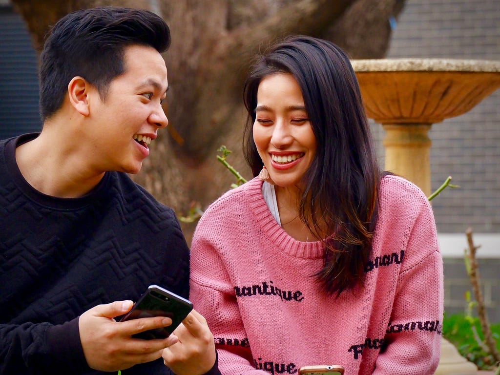 Two people wearing knitted jumpers are laughing while looking at their smartphones.