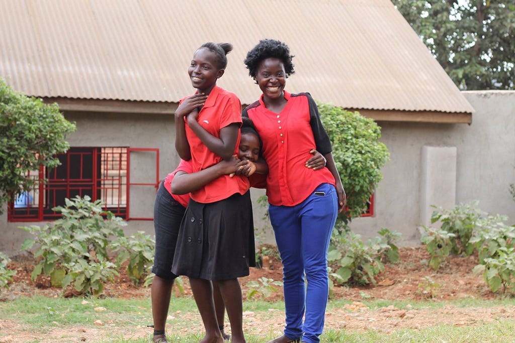 Three young Black women in red shirts embrace in front of a house.