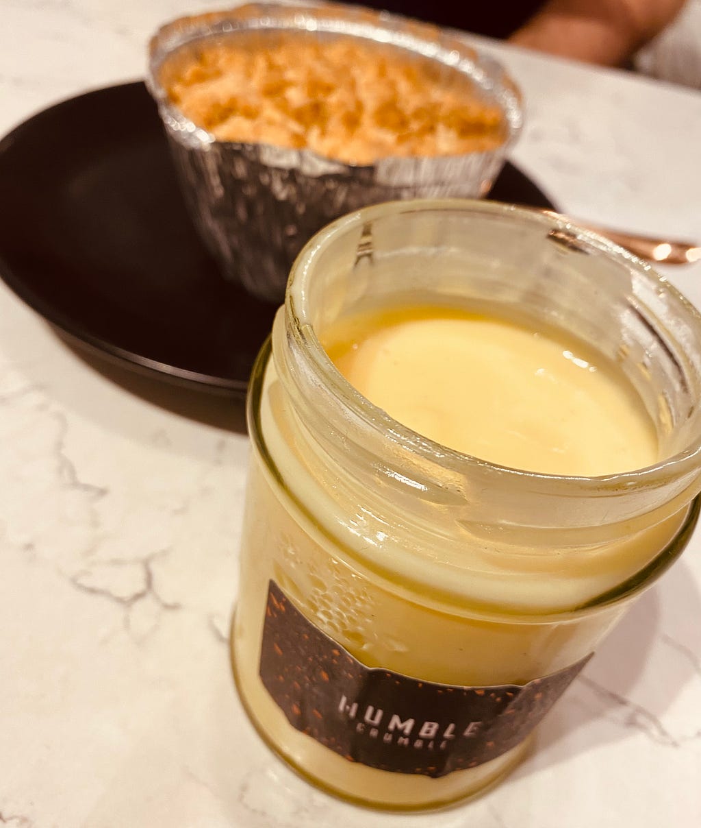 A glass jar that contains custard on it on the front and faintly visible on the back a crumble served on a tin foil plate.