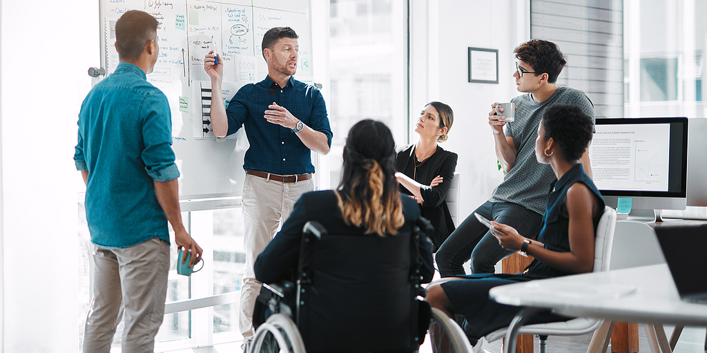 A group of coworkers casually gather around a whiteboard to discuss a kanban board.