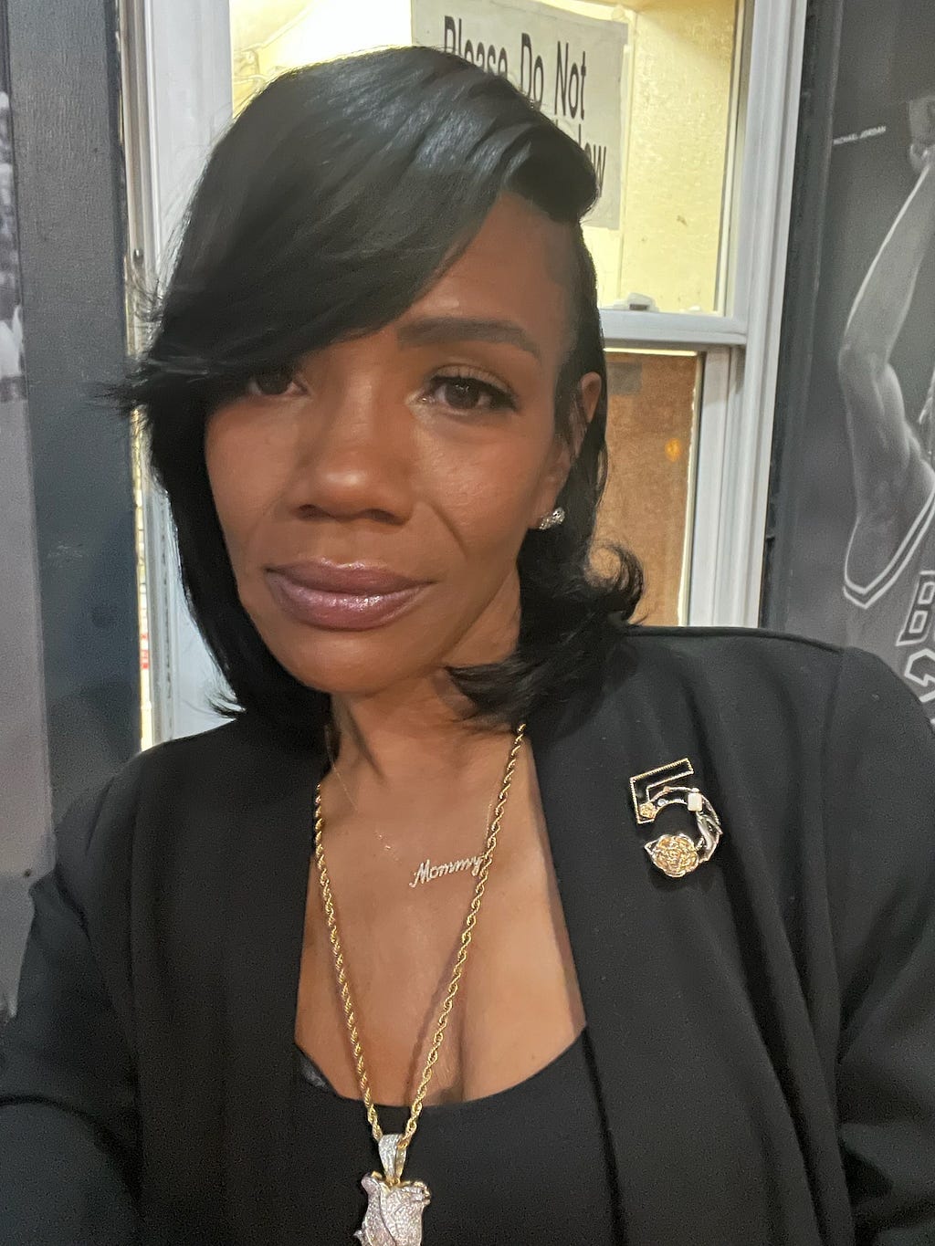 A Black woman with a sleek side parted bob haircut wearing a stylish black suit with gold jewelry takes a selfie.
