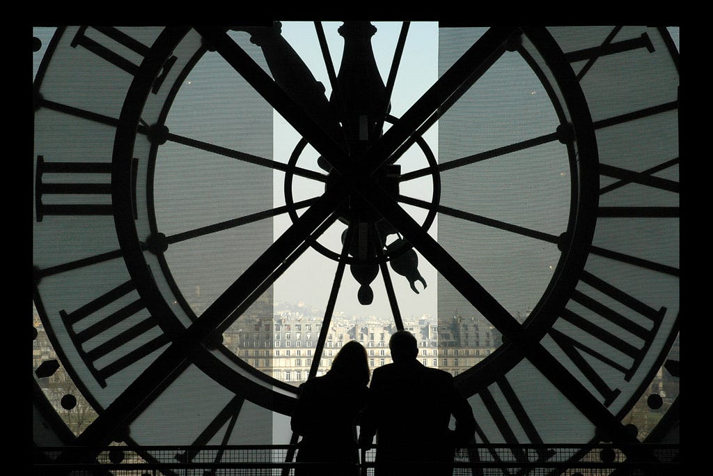 Silhouettes of two people in front of the inside of a large, clear clock overlooking a city.