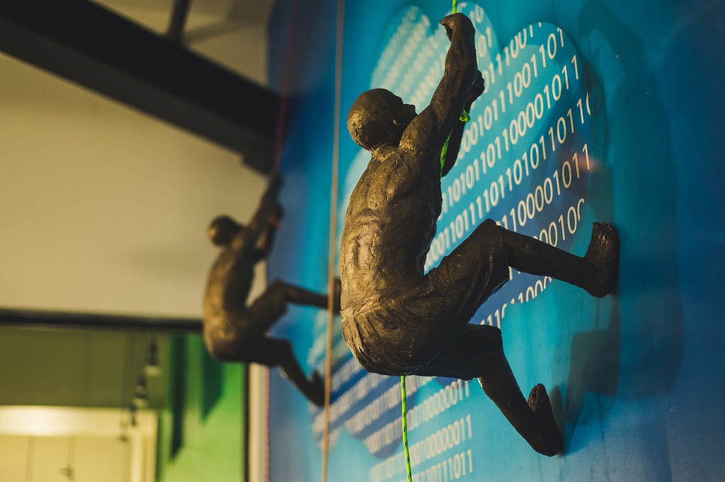 Two bronze figurine men climbing a wall with binary code printed on it.