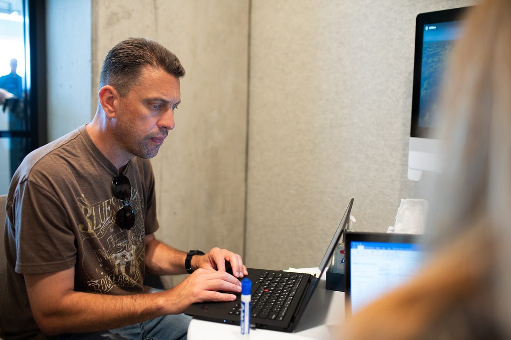 An Expedia Group employee in a brown T-shirt works on their laptop next to a colleague.