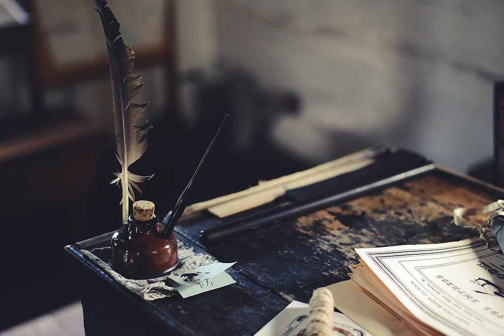 A dark wooden desk has a stack of paper and a pot of ink with a quill and fountain pen in it.