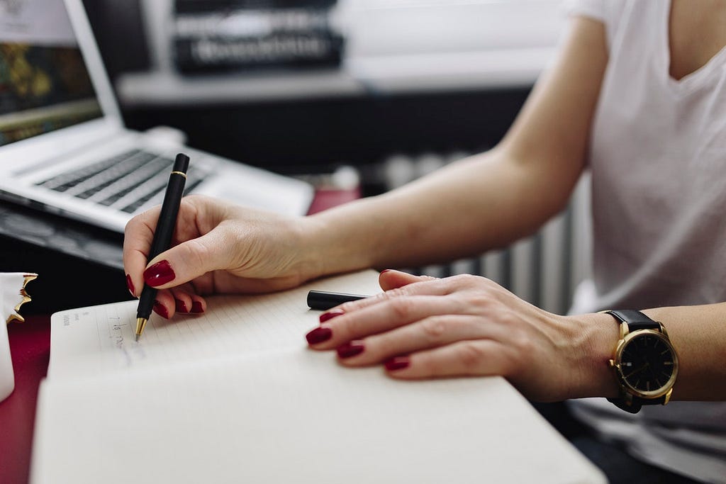 Arms and torso of woman writing with pen, laptop in background