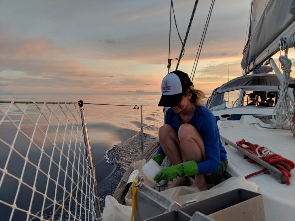 Woman in cap squats on boat around sunset. She is wearing green globes and holding a plastic item.