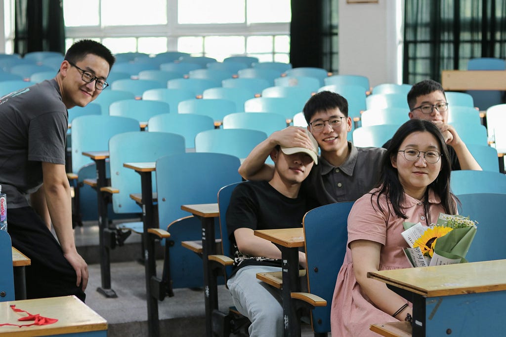 The image shows a bright classroom with four cheerful individuals among rows of teal chairs and desks. From left to right, there’s a standing man in a grey t-shirt, a seated man in a black t-shirt playfully covering his face, another standing man in a light grey shirt, and a seated woman in a pink shirt holding a bouquet of flowers. The mood is casual and friendly.