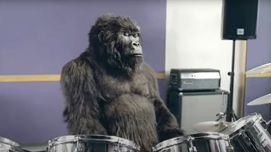 Gorilla sitting at a drum kit, eyes closed, sitting in a purple music studio.