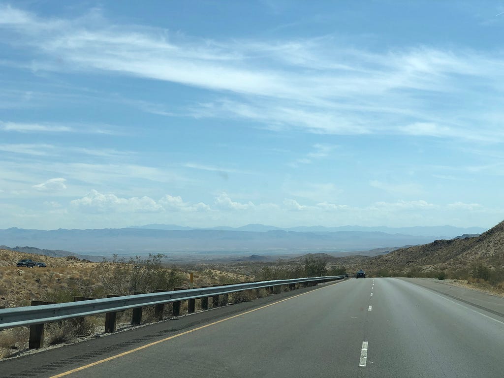 Picture taken from a car on a highway on a hillside, showing an expansive view of a desert basin and far-off mountains.