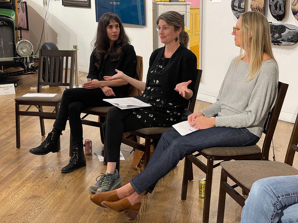 Three women sit in chairs talking to a group. On the left is a woman with long curly black hair and bangs, in the middle is a woman speaking with her arms held wide. She has grey hair pulled into a pony tail and glasses. On the right is a woman with long blonde hair and glasses.