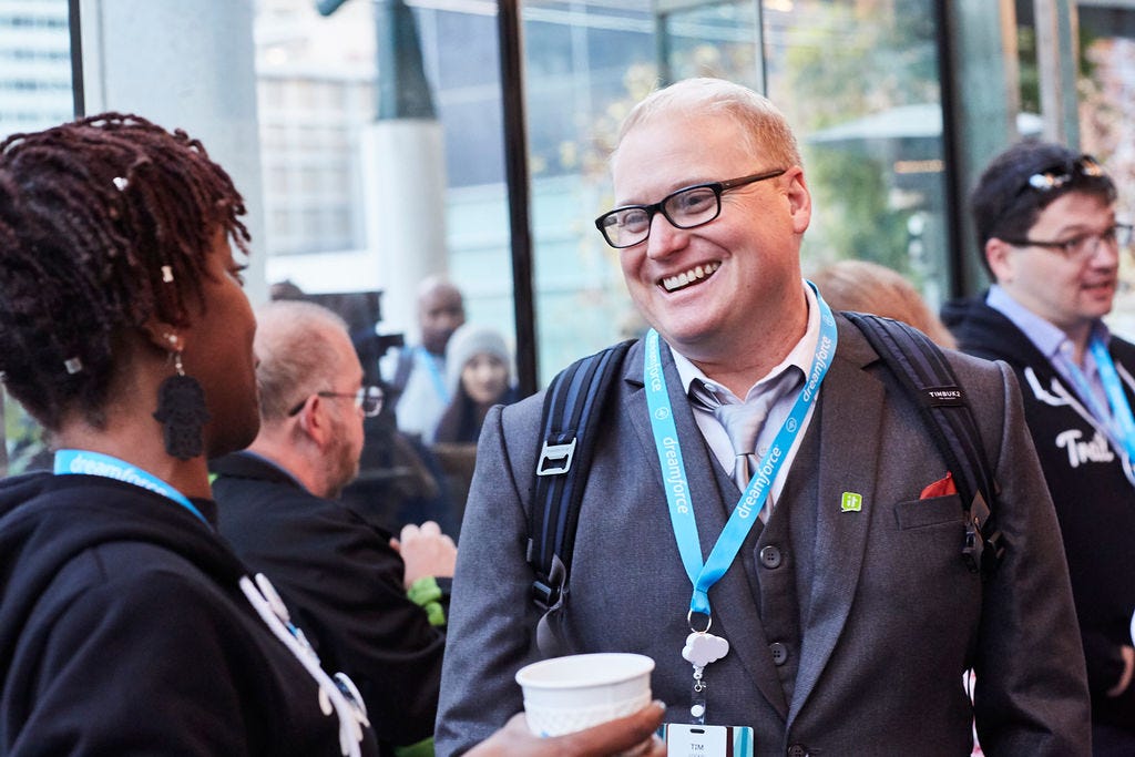 A smiling man with glasses and lanyard speaks with a woman holding a beverage at a Salesforce event.