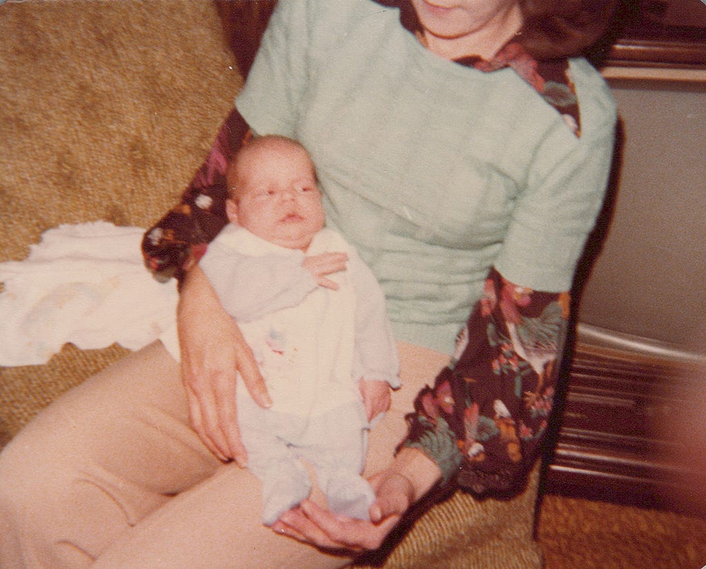 Newborn baby in sleeper with uncomfortable facial expression being held by adoptive mother with 70s style clothing and furninshings