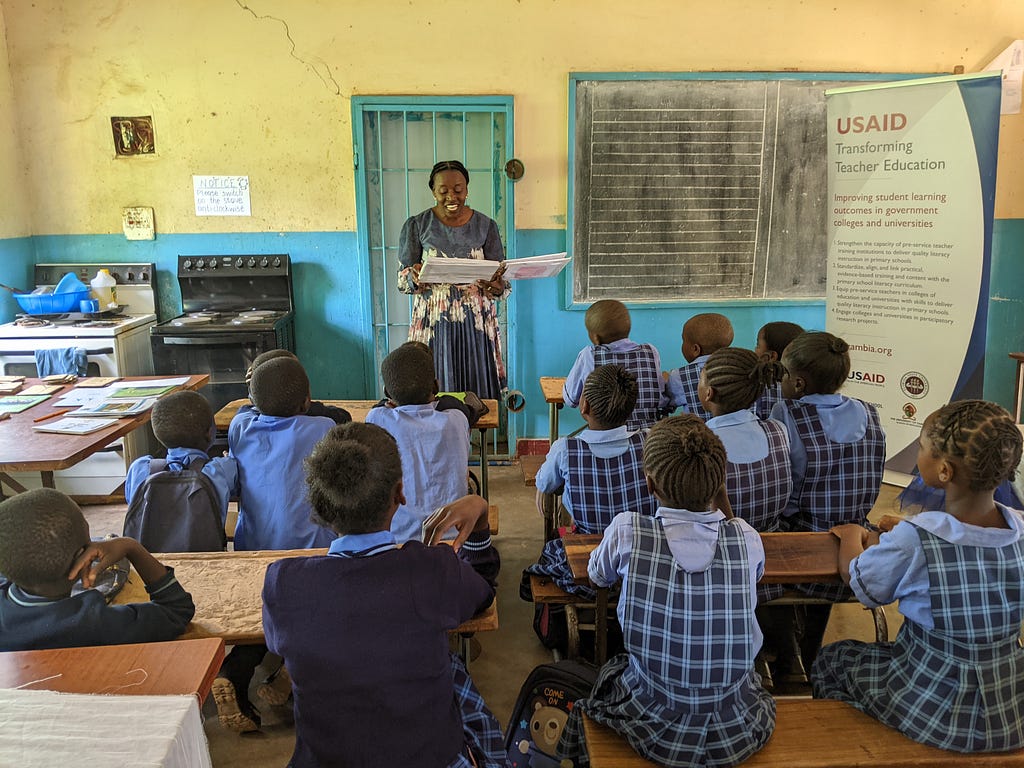 A student teacher stands at the head of a classroom reading from a book with more than a dozen students sitting at desk in front of her.