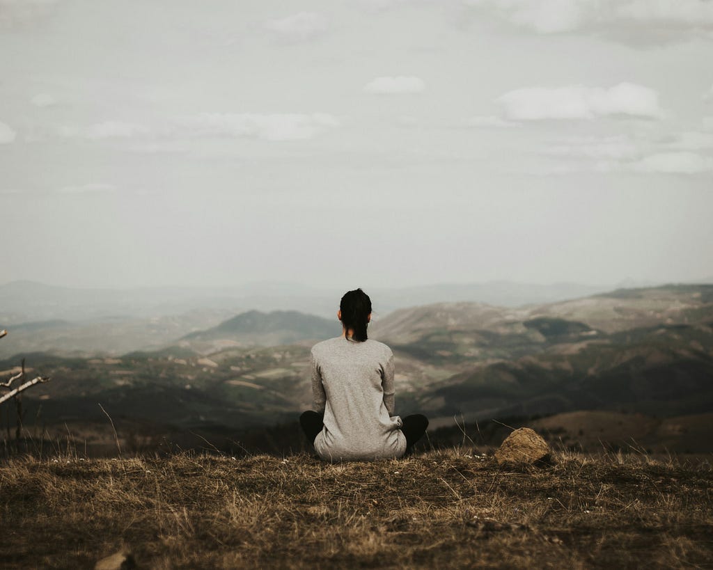 A woman sits on the edge of a hill, gazing upon a vast valley ahead of her. She is contemplative